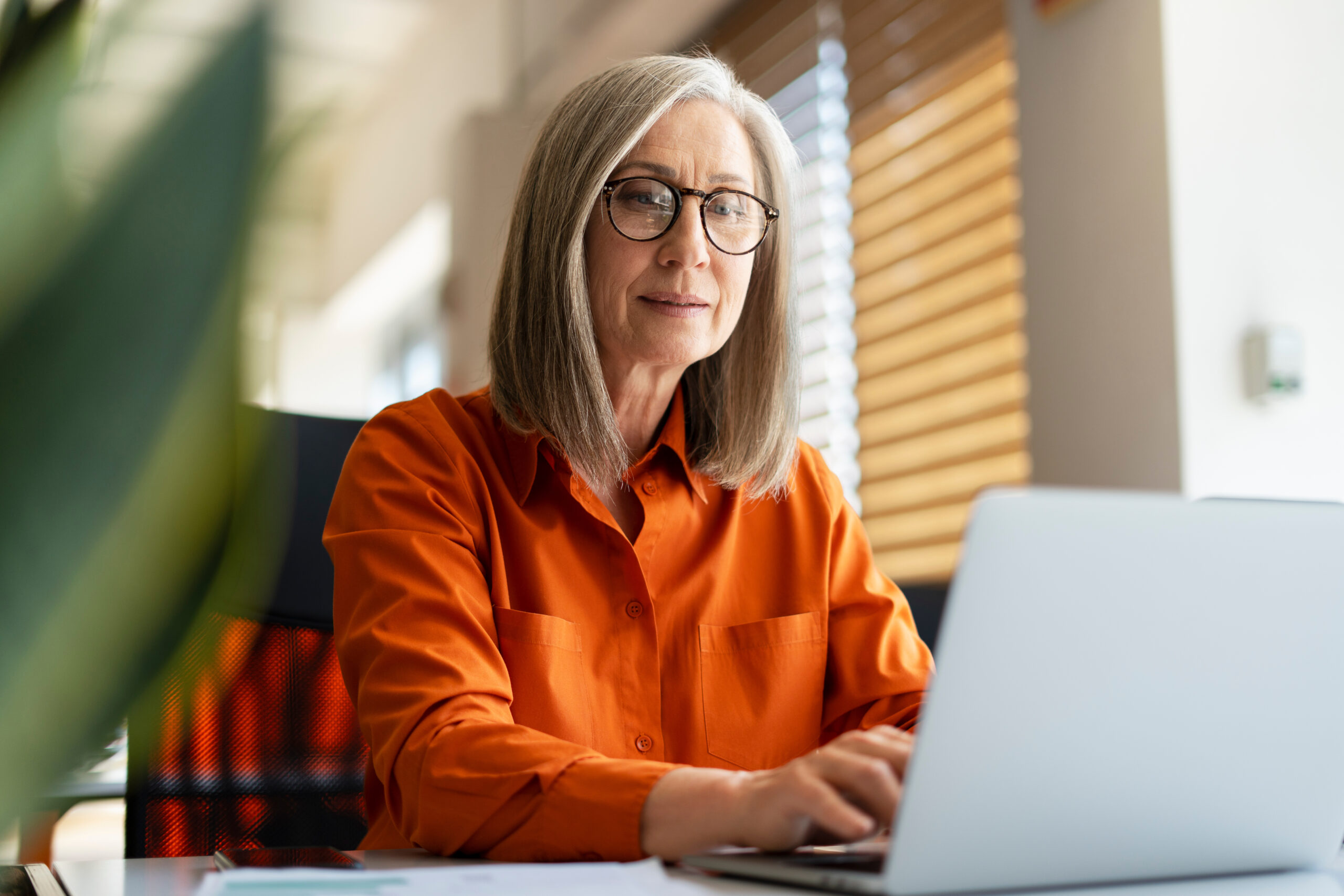 Portrait of confident serious mature businesswoman wearing stylish eyeglasses, orange shirt using laptop working online in modern office. Successful manager planning project, typing on keyboard Community Care Corps Supports Volunteer Caregivers